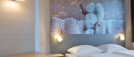 A hotel room with a bed, a nightstand, a wall-mounted lamp, and a large cotton-themed artwork.