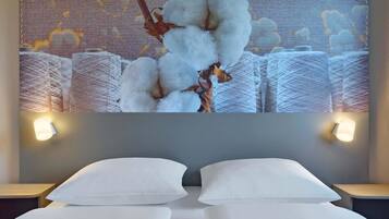 Une chambre d’hôtel comprenant un lit, deux oreillers, une œuvre d’art en coton représentant des capsules de coton, et deux appliques murales.