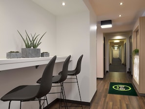 Zona de recepción de hotel con un mostrador blanco, tres taburetes negros y plantas en macetas sobre el mostrador.