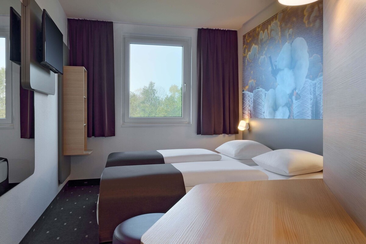 A hotel room with a bed, two bedside tables, a TV mounted on the wall, and a window with curtains.