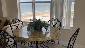 A round dining table with chairs, a potted plant, and a view of the beach through large windows.