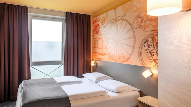A hotel room with a bed, a window with curtains, a wall with a decorative pattern, and a bedside lamp. - B&B Hotel Schweinfurt-Süd (Schweinfurt)