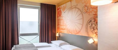 A hotel room with a bed, a window with curtains, a wall with a decorative pattern, and a bedside lamp.
