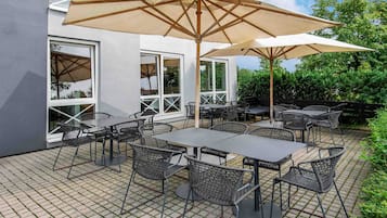 Un patio al aire libre con mesas y sillas, grandes sombrillas y un edificio con amplias ventanas.