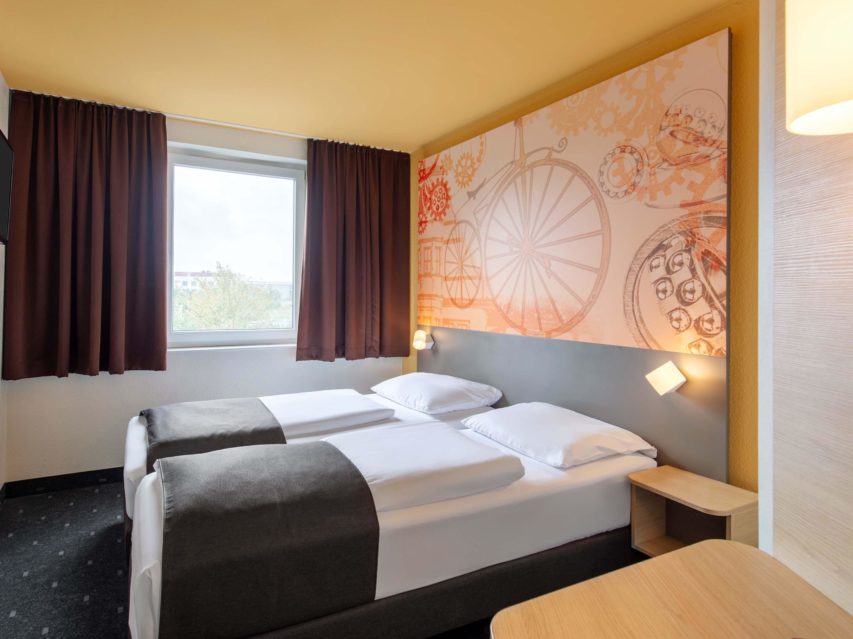 Habitación de hotel con dos camas, una ventana con cortinas, un mural temático de bicicletas en la pared y una mesita de noche con lámpara.