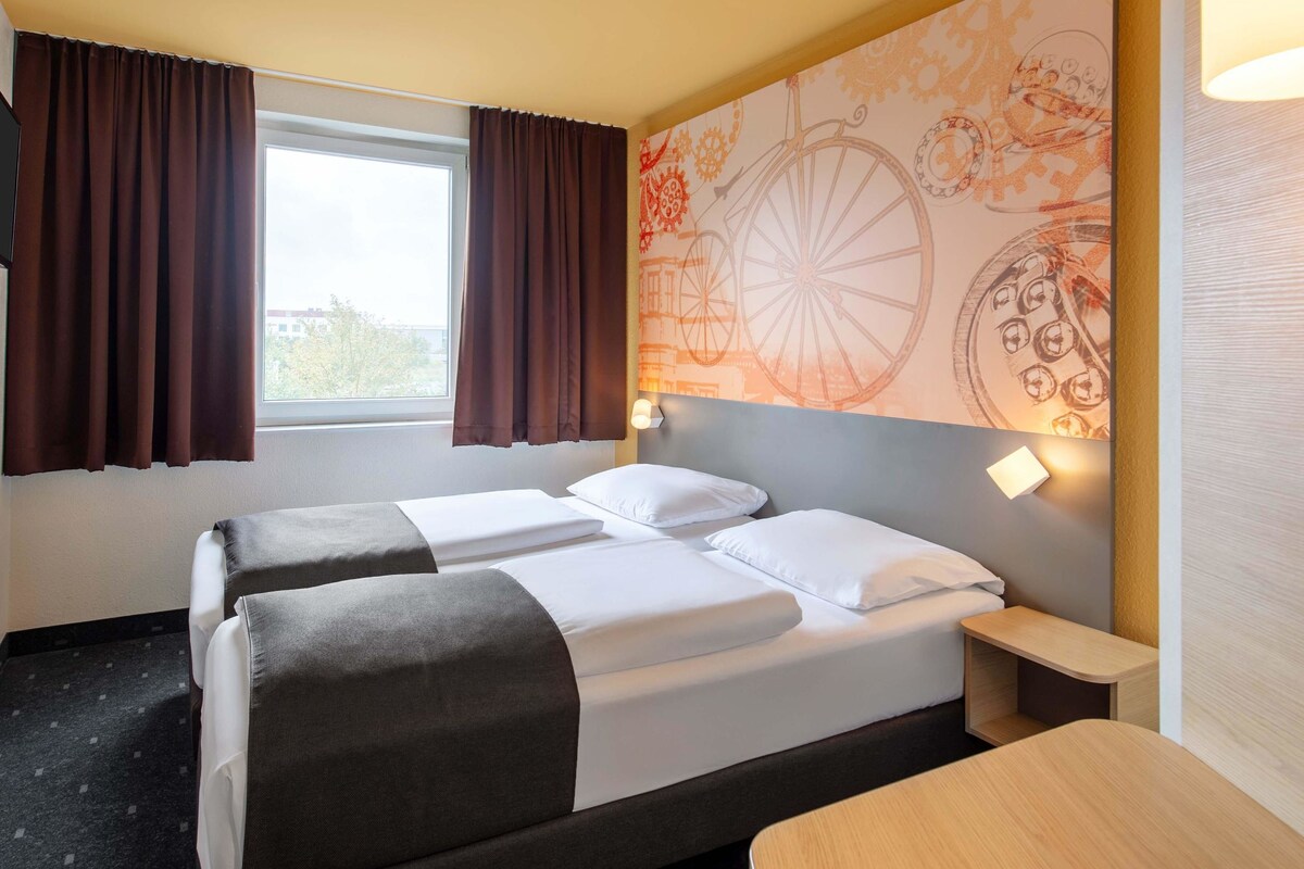 Une chambre d’hôtel avec deux lits, une fenêtre avec des rideaux, un mur orné d’une fresque sur le thème du vélo, et une table de chevet avec une lampe.