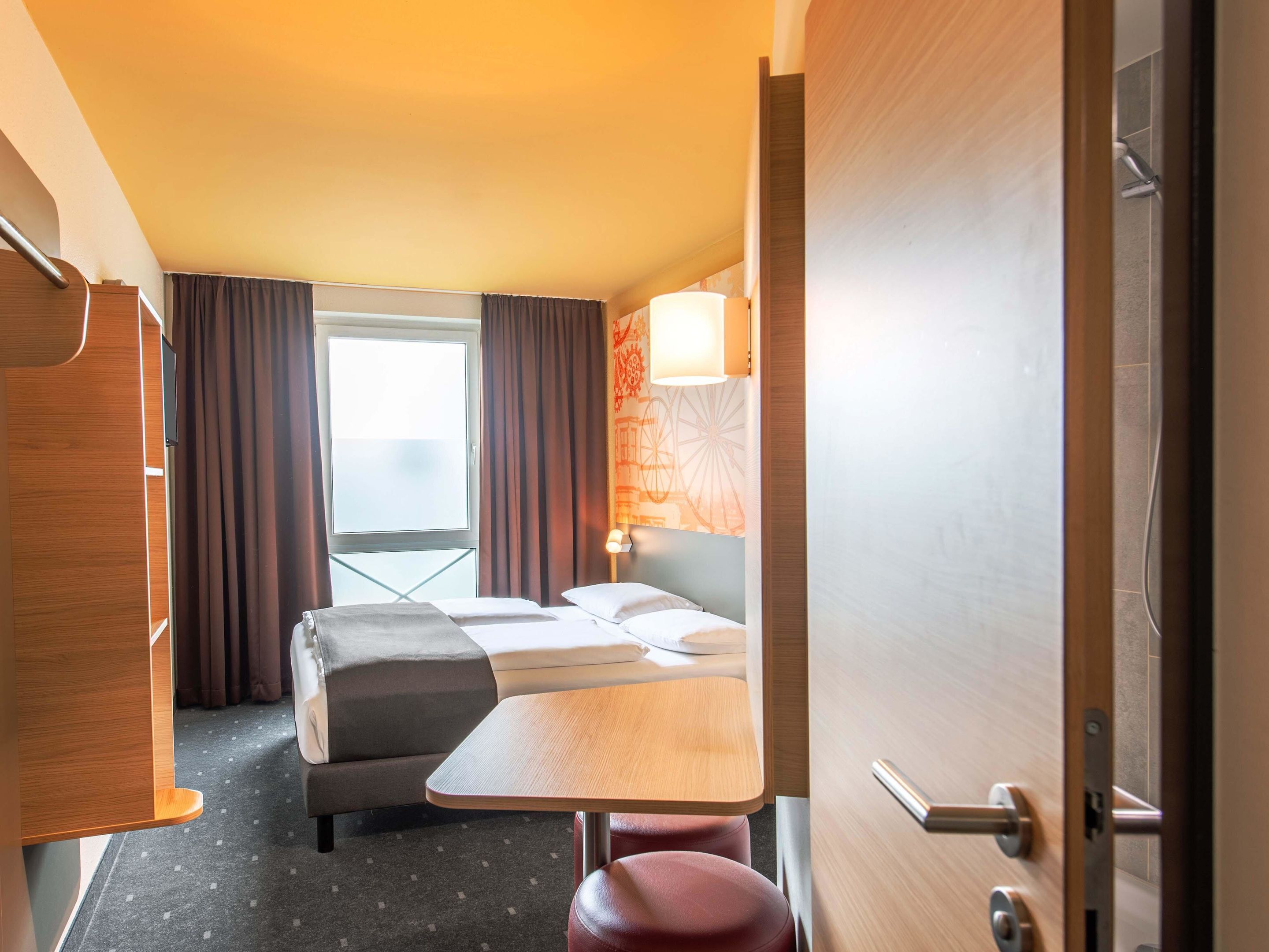 Ein Hotelzimmer mit Bett, kleinem Tisch, Stuhl, Schreibtisch und einem Fenster mit Vorhängen.