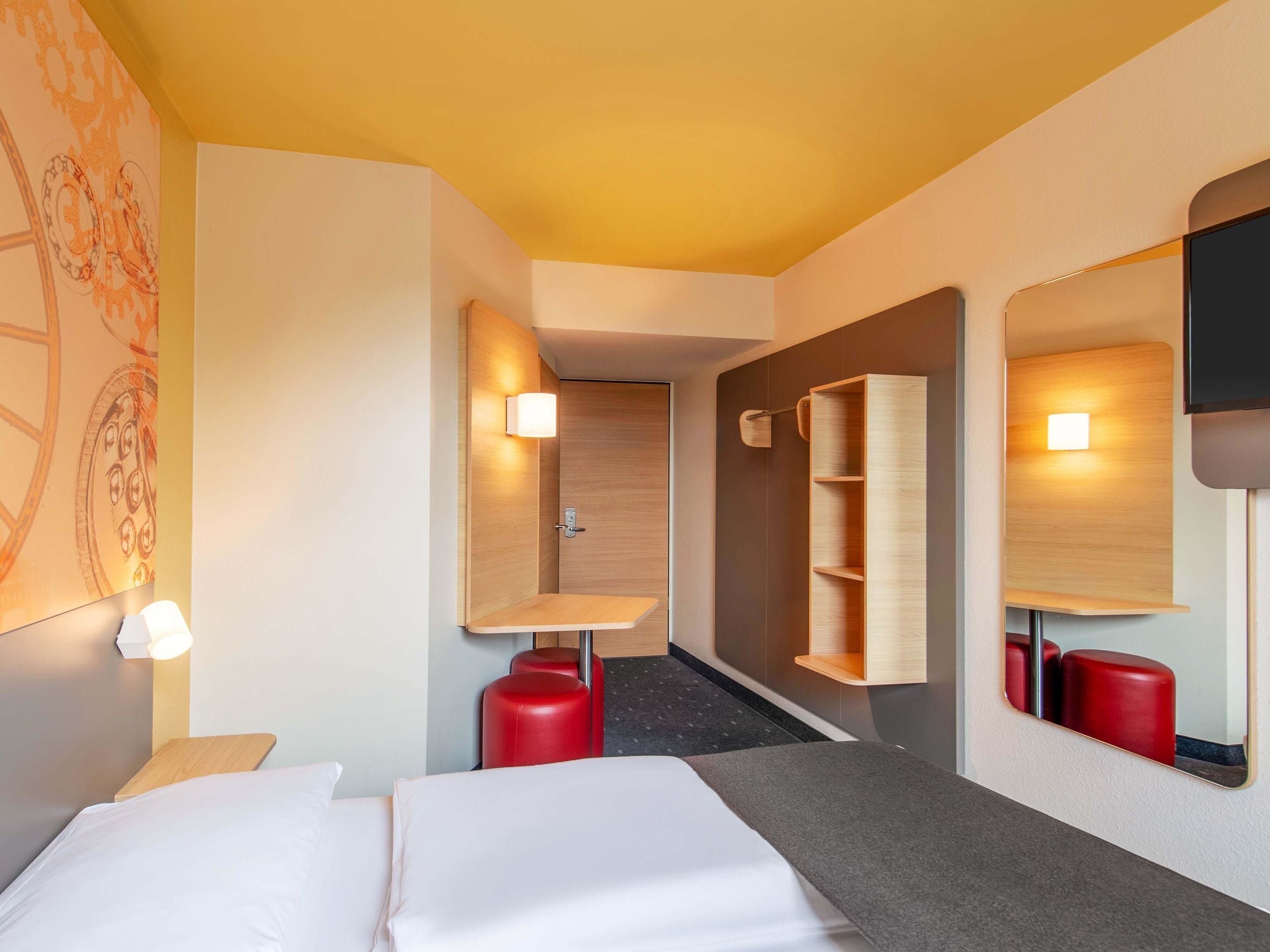 A hotel room with a bed, a wooden wardrobe, a mirror, and a red stool.
