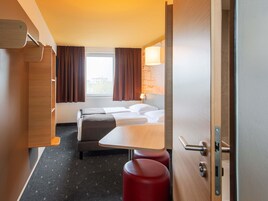Une chambre d’hôtel avec un lit, une petite table et un tabouret rouge. Il y a une fenêtre avec des rideaux et une lampe fixée au mur.