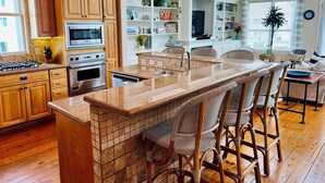 A modern kitchen with wooden cabinets, a built-in island with bar stools, a large window, and a dining area with wicker chairs. - Booking NOW for fall! 5 min walk to beach! Wrap @decks. Gulf Views! (Galveston)