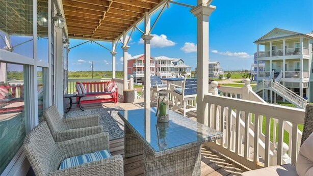 A covered outdoor area with wicker furniture, a glass table, and a view of multiple buildings. - Booking NOW for fall! 5 min walk to beach! Wrap @decks. Gulf Views! (Galveston)