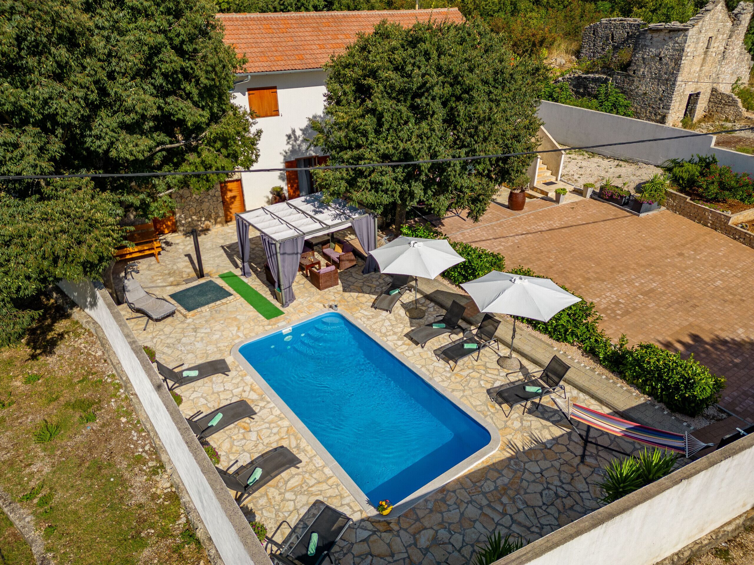 Ovenfra ses et område med swimmingpool, liggestole, parasoller og et overdækket opholdsområde.