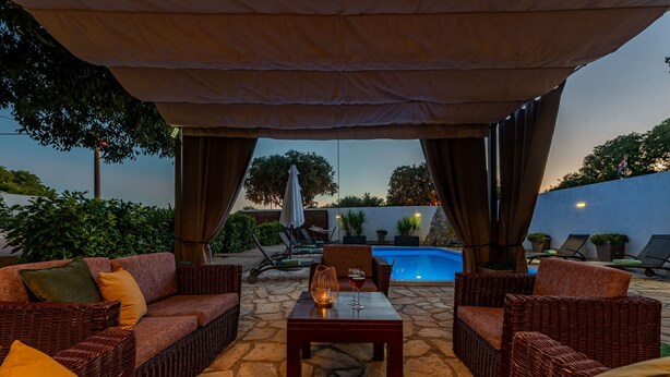 An outdoor seating area with wicker furniture, a table with a candle, and a pool in the background. - Villa Natura (Benkovac)