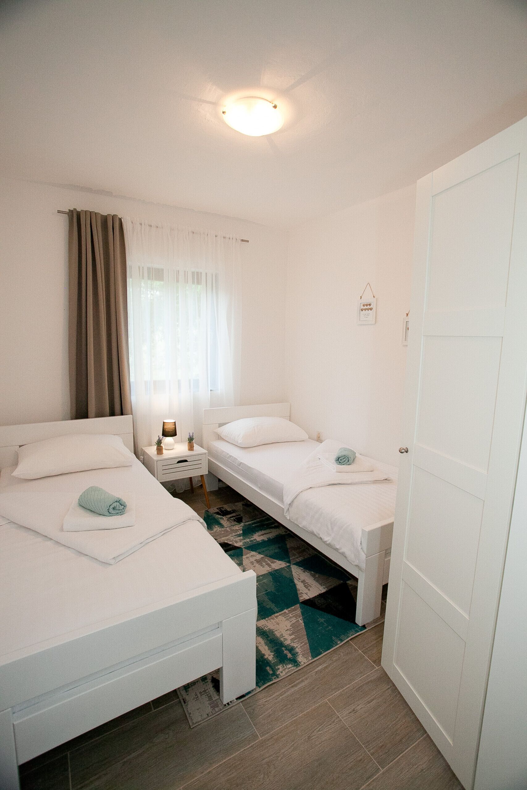 A hotel room with two single beds, a small bedside table with a lamp, a window with curtains, and a door.