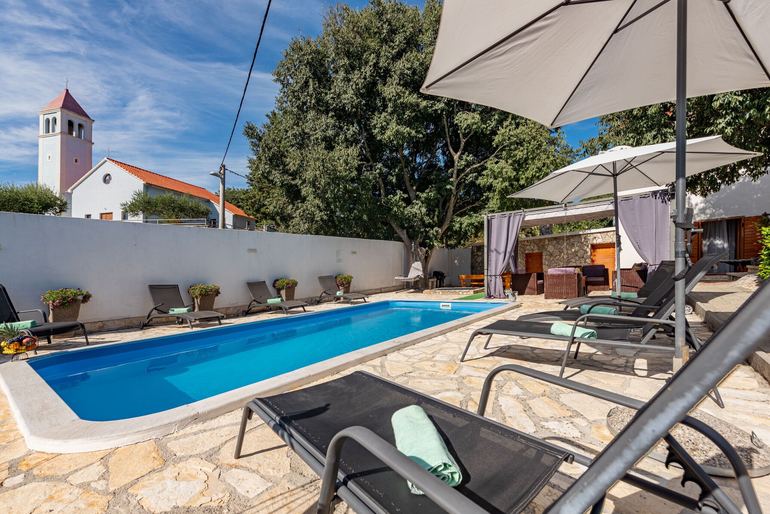 A swimming pool with lounge chairs and umbrellas, a stone wall, and a building with a red roof and a bell tower.