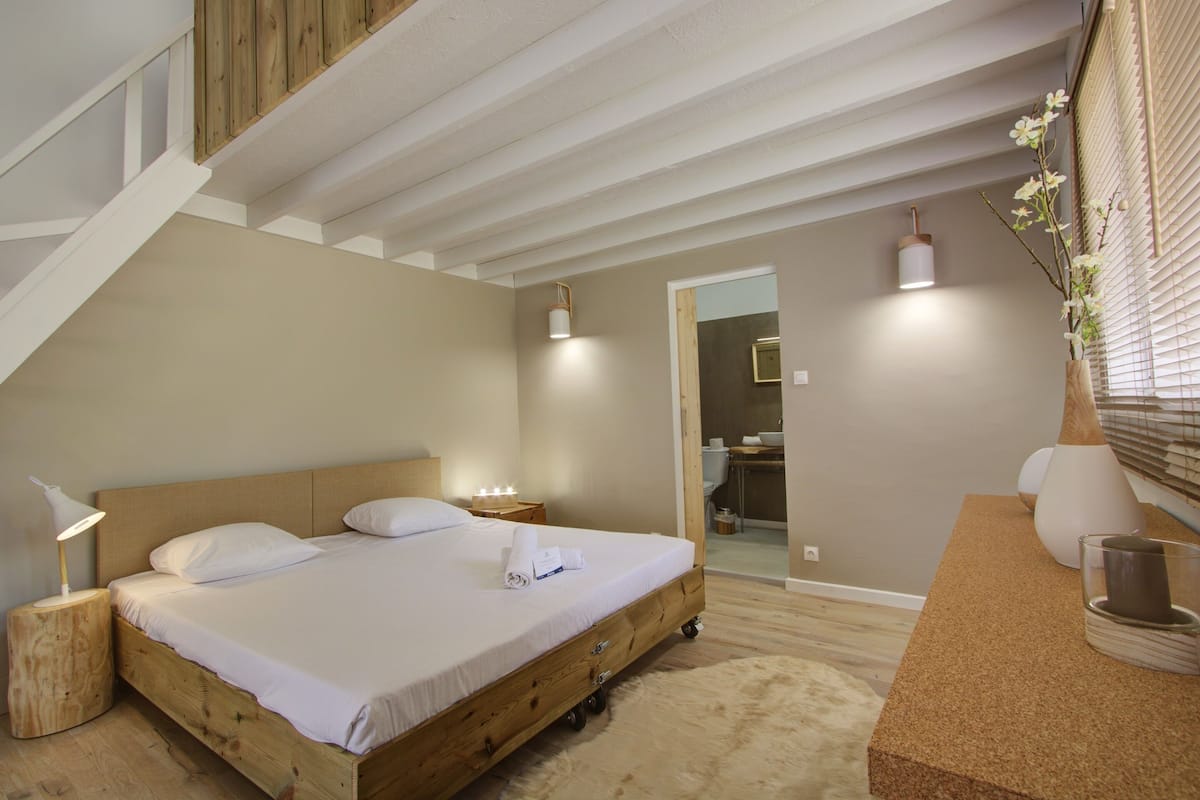 Un dormitorio con una cama de madera, ropa de cama blanca, una mesita de noche con una lámpara y una pequeña mesa con un florero.
