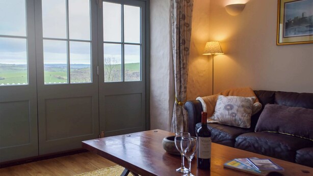 A room with a wooden table, a bottle of wine, two glasses, a sofa, a lamp, and a view through a window. - 2 bedroom accommodation in Dwrbach, near Fishguard (Dwrbach, near Fishguard)