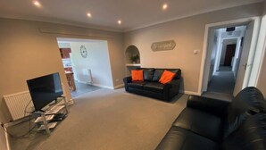 A living room with a black leather sofa, a flat-screen TV, and a 'Be our guest' sign on the wall. - 4 bedroom accommodation in Newton Stewart (Newton Stewart)