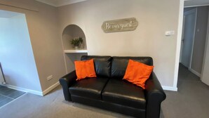 A black leather sofa with orange cushions, a 'Be our guest' sign on the wall, and a small plant on a shelf. - 4 bedroom accommodation in Newton Stewart (Newton Stewart)
