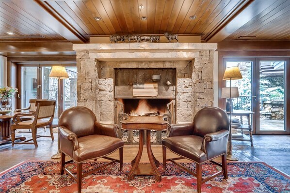 A stone fireplace with a fire, two leather chairs, a wooden table, and a patterned rug in a room with wooden ceilings and large windows. - 1-#42 4BD One Willow Bridge Road (Vail)