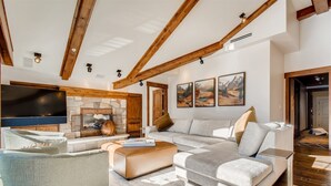 A modern living room with a stone fireplace, wooden beams, a large flat-screen TV, and a sofa set. - 1-#42 4BD One Willow Bridge Road (Vail)