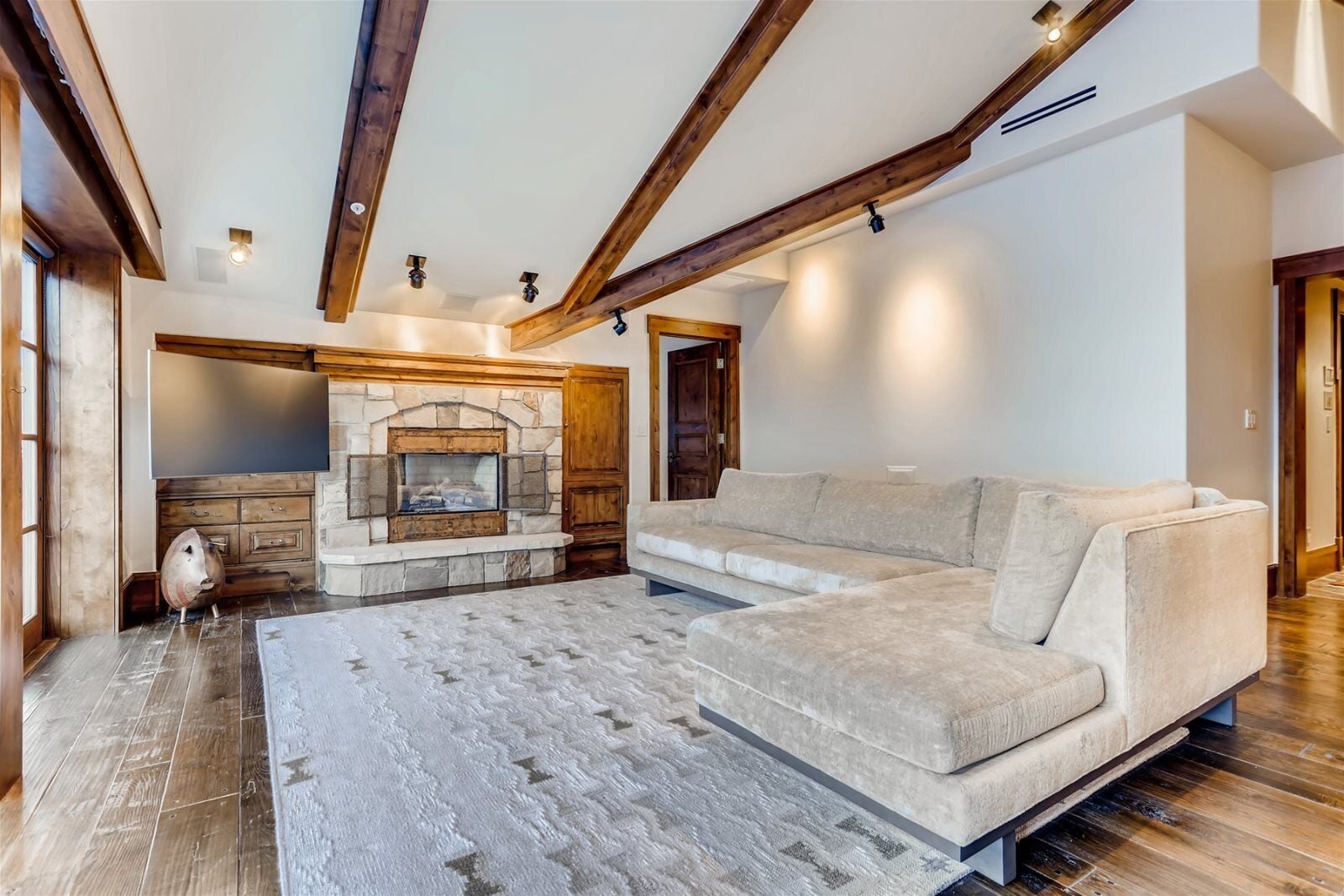 A living room with a stone fireplace, wooden beams, a beige sofa, and a patterned rug.