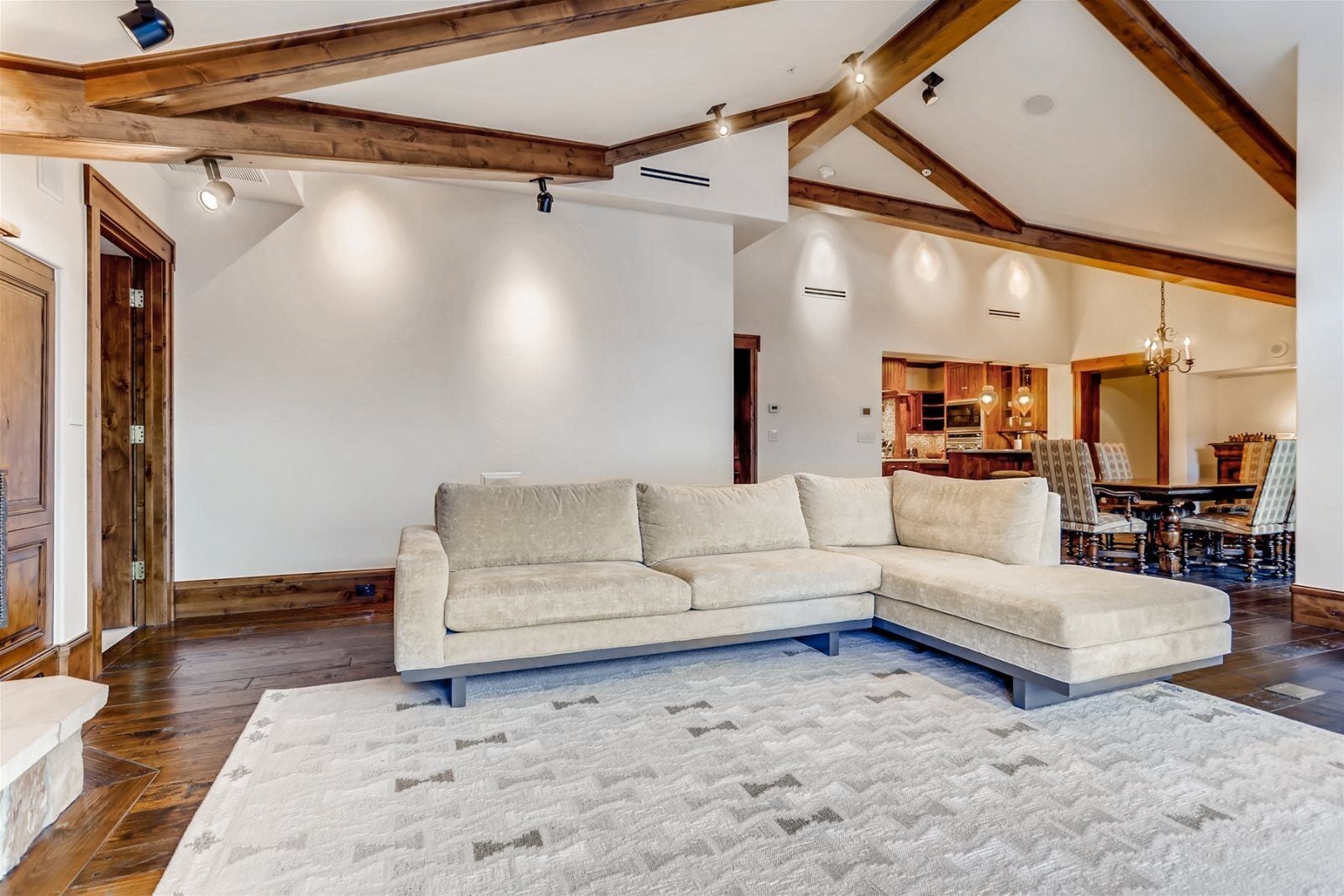 Una sala de estar con un sofá seccional color beige, vigas de madera y un comedor con una araña.