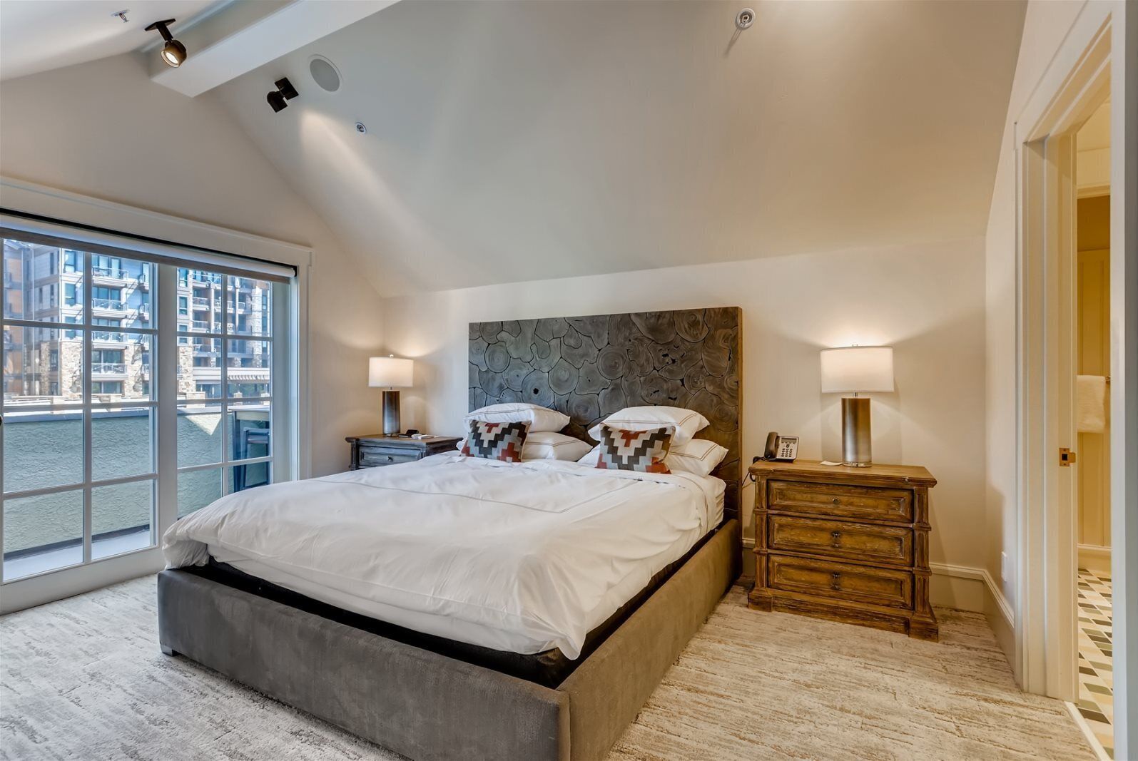 A modern bedroom with a large bed, a stone headboard, and a view of buildings through a large window.