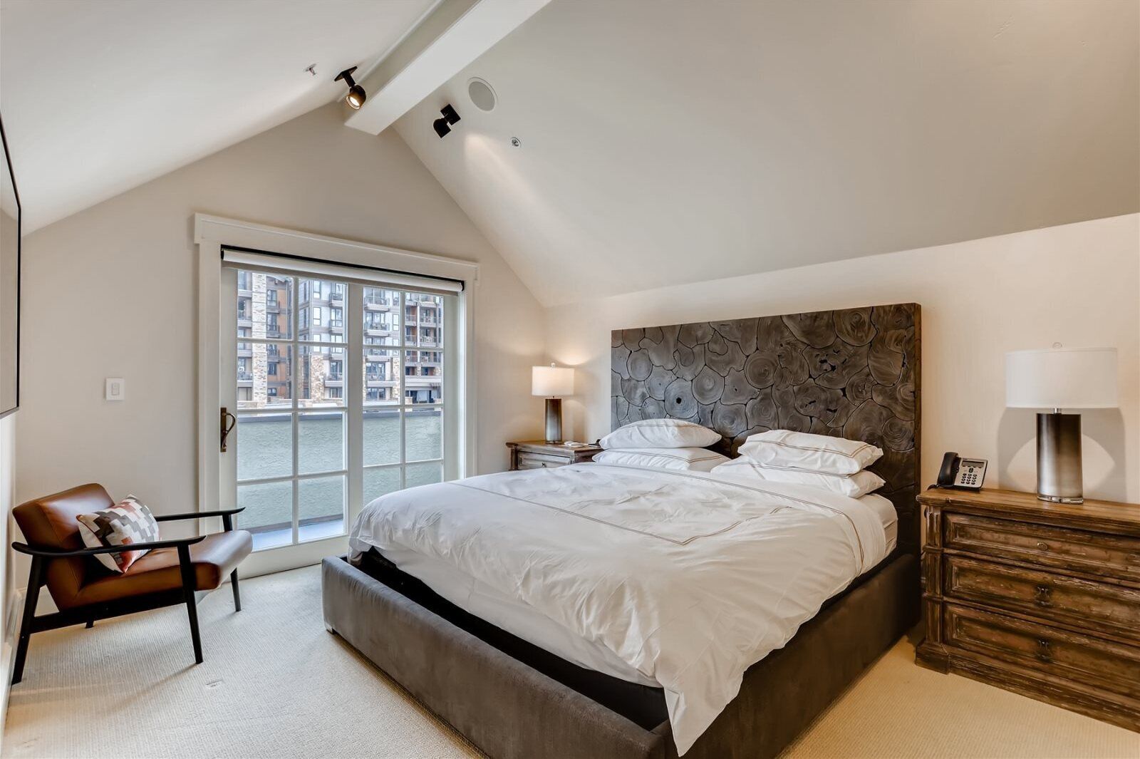 A bedroom with a large bed, a chair, a nightstand, a lamp, and a view of a building through a window.