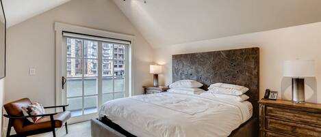 A bedroom with a large bed, a chair, a nightstand, a lamp, and a view of a building through a window.