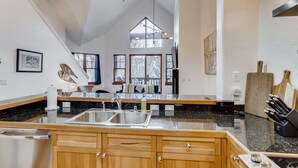 A modern kitchen with granite countertops, wooden cabinets, a large island with a sink, and a dining area with a view of the outdoors. - 1-brookside 307 (Avon)
