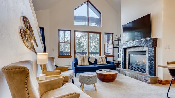 A spacious living room with a high ceiling, a fireplace, and large windows offering a view of the outdoors. - 1-brookside 307 (Avon)
