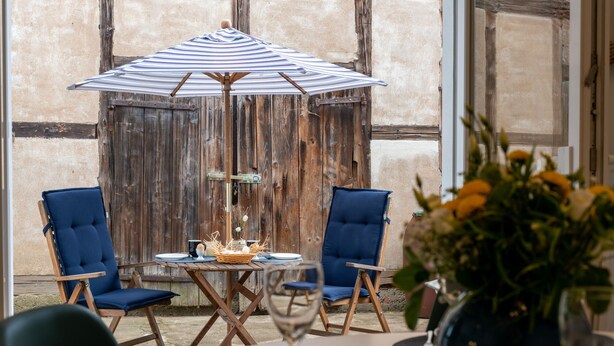 A dining area with a wooden table, two blue cushioned chairs, a striped umbrella, and a vase with flowers.