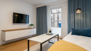 A modern hotel room with a bed, a bedside table, a TV mounted on the wall, a window with curtains, and a view of the outside.