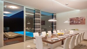 A modern dining room with a long table, white chairs, and a view of a pool area. - Luxury Beachfront Villa Blue Star Brac with Pool (Milna)