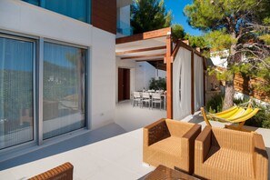 A modern outdoor patio with wicker furniture, a hammock, and a dining area visible through a glass door.