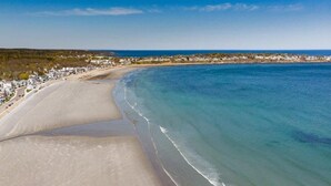 Eine Vogelperspektive zeigt einen Strand mit einer geschwungenen Küstenlinie, klares blaues Meer und eine Wohngegend im Hintergrund.