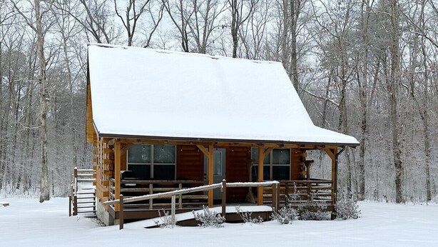 Eine Holzhütte mit schneebedecktem Dach und einer Veranda, umgeben von einem verschneiten Wald.
