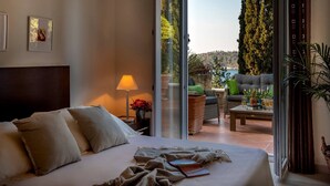 A hotel room with a bed, a nightstand, a lamp, a view of the outdoors, and a balcony with seating and plants.