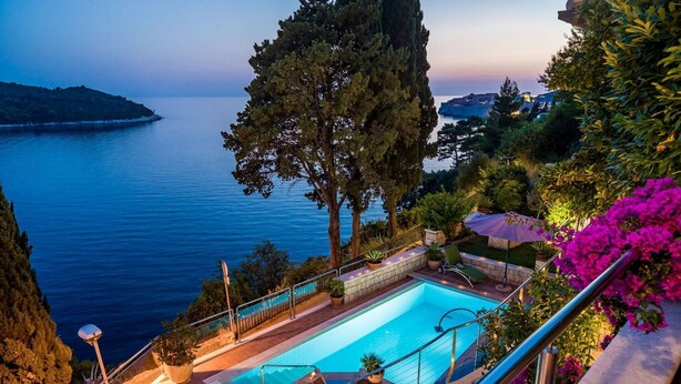 A pool with a view of the sea, surrounded by greenery and stairs leading down to the water.