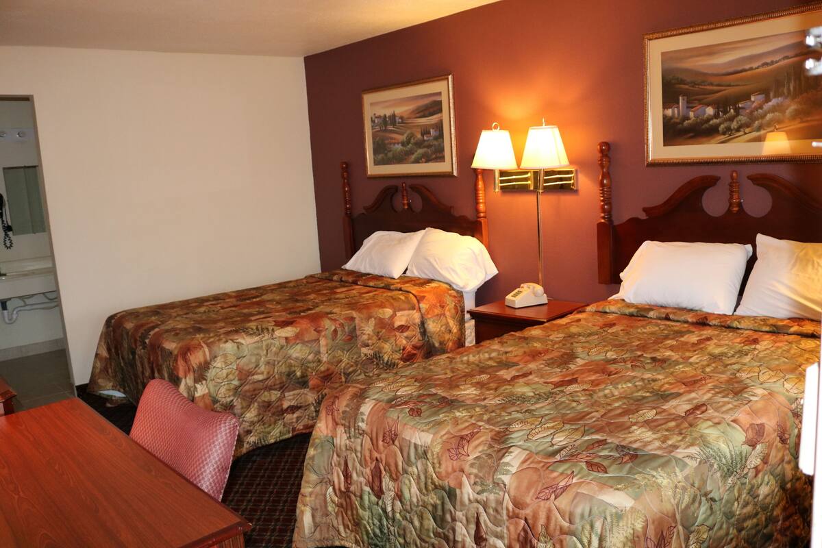 A hotel room with two beds, a desk, and a chair. There are two wall-mounted lamps and a painting above the beds.