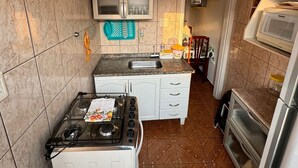 A compact kitchen with a gas stove, microwave, and oven. There is a cabinet with glass doors above the sink, and a small countertop with various items. - Apartment 100 meters from Praia Grande – Vila Tupi (Praia Grande)