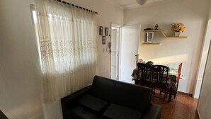 A living room with a black sofa, a ceiling fan, a dining area with a table and chairs, and a kitchen visible through an open door. - Apartment 100 meters from Praia Grande – Vila Tupi (Praia Grande)