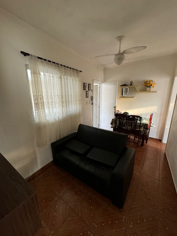 Ein Wohnzimmer mit schwarzem Sofa, Deckenventilator, Essbereich mit Tisch und Stühlen sowie einer durchgehenden Tür sichtbaren Küche.