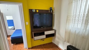 A room with a TV mounted on a wooden panel, a black leather sofa, and a bed with a blue mattress. - Apartment 100 meters from Praia Grande – Vila Tupi (Praia Grande)