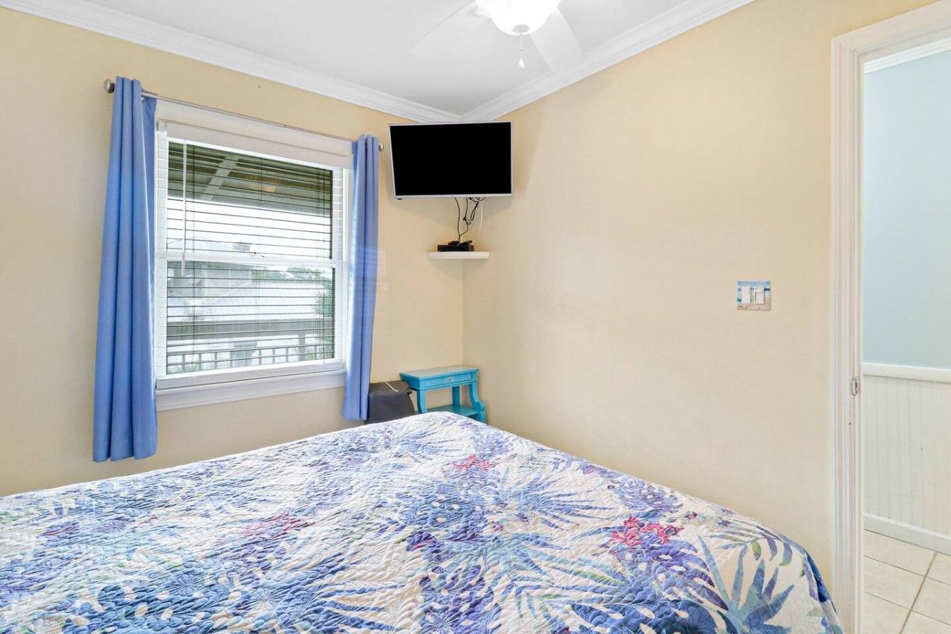 Un dormitorio con cama, televisor montado en la pared, ventana con cortinas y una mesita pequeña.