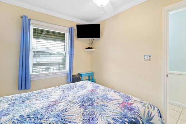 A bedroom with a bed, a television mounted on the wall, a window with curtains, and a small table.
