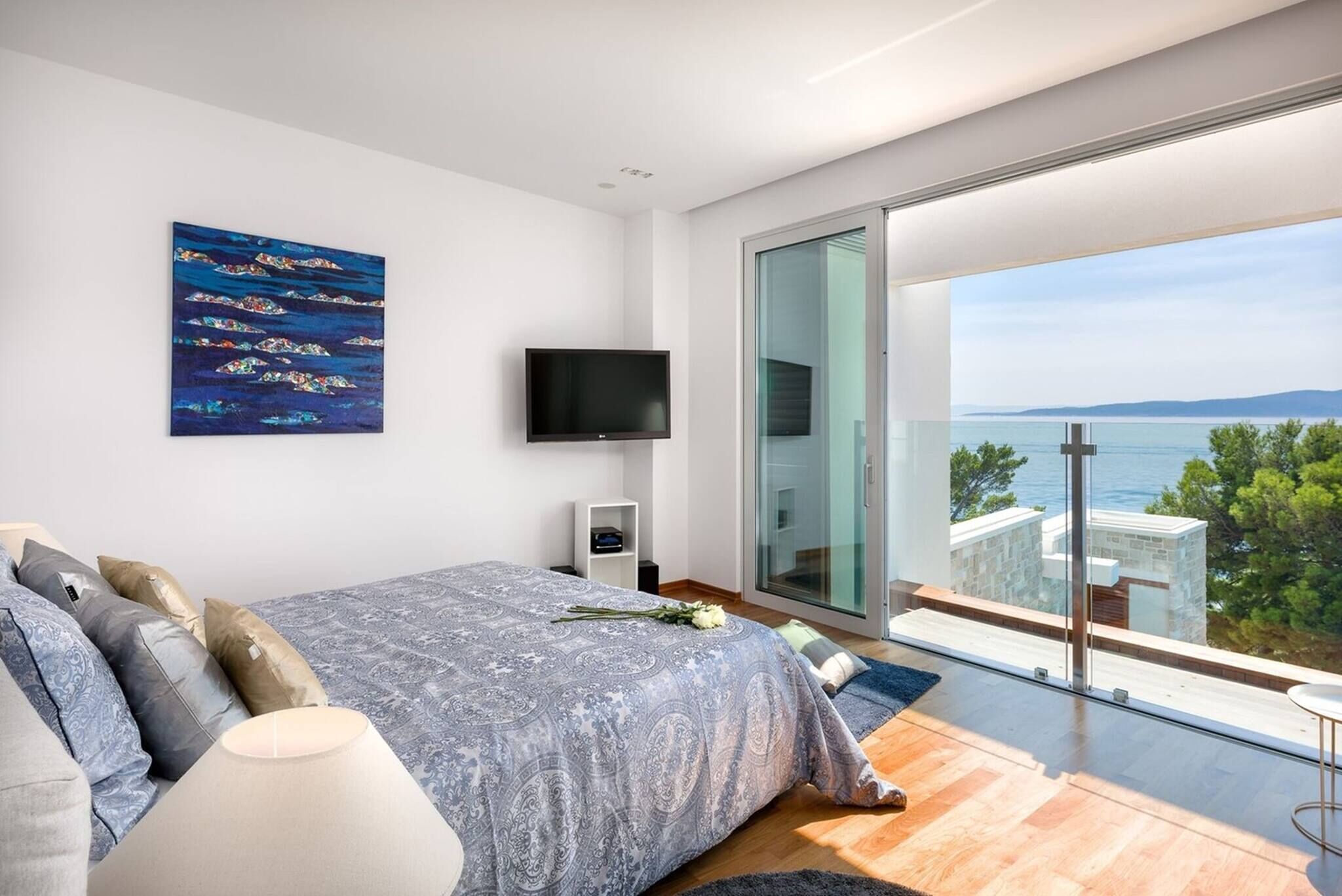 Ein modernes Schlafzimmer mit einem großen Bett, einem Flachbildfernseher, einem Gemälde und einem Balkon mit Meerblick.