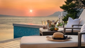 A sunset view from a balcony with a pool, wicker furniture, and a wicker basket with fruits and glasses.