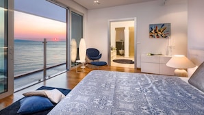 A bedroom with a large bed, a chair, a desk, and a view of the sea at sunset.